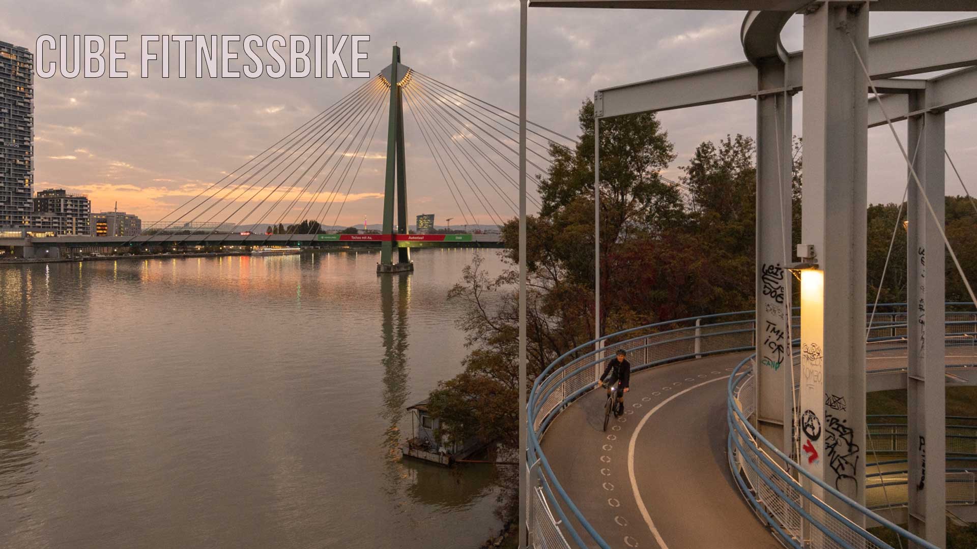 Si vede una città sul fiume, in primo piano una ciclista che pedala sulla pista ciclabile al tramonto. L'immagine è intitolata Cube Fitnessbike.