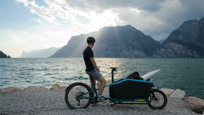 Un uomo è in piedi con la sua bicicletta da carico, in riva al lago. Guarda le montagne sullo sfondo.