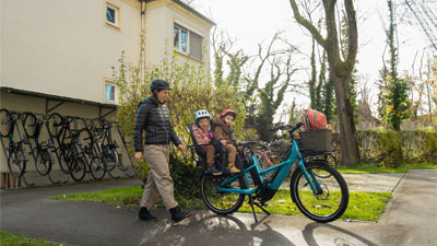 Si vede una madre che corre verso la sua bicicletta da carico blu. Sul portapacchi sono seduti due bambini piccoli, la scena si svolge in un quartiere residenziale.