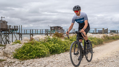 Si vede un uomo in tenuta da ciclista. Sta pedalando con la sua bici gravel su una strada lungo la spiaggia.