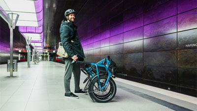 Si vede un uomo in piedi in una stazione della metropolitana. Davanti a lui c'è una bicicletta pieghevole blu.