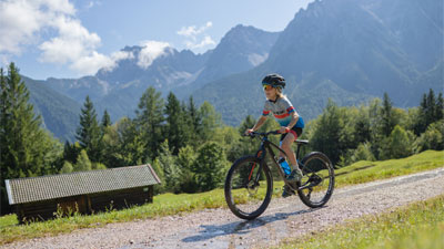 Un ragazzo va in mountain bike su una strada sterrata attraverso un paesaggio montuoso. Sullo sfondo si vede una capanna. 
