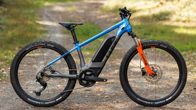 Si vede una bicicletta elettrica per bambini Cube grigia con dettagli blu e arancioni. Si trova su un sentiero nel bosco.